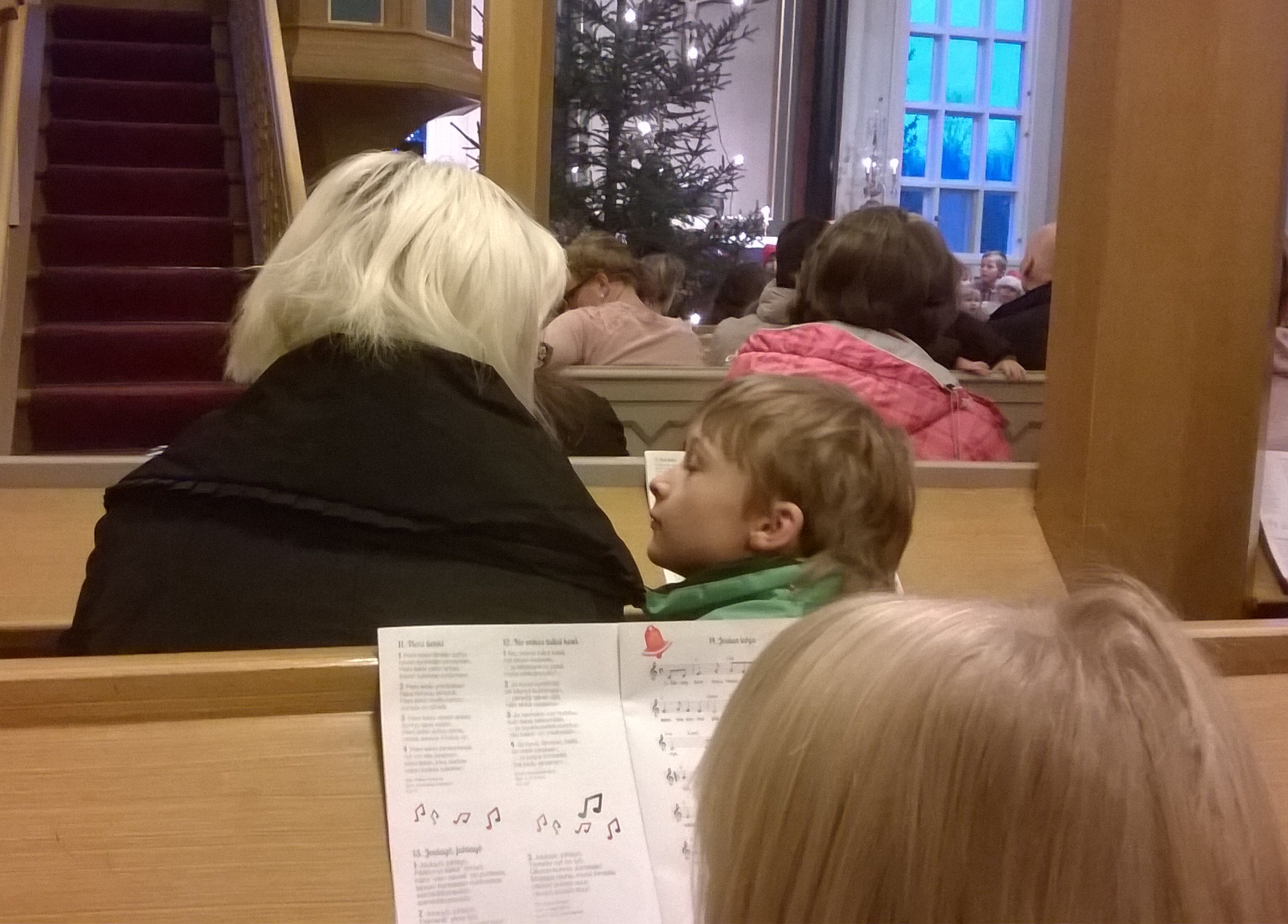 Kuva kauneimpien joululaulujen laulamisesta kirkossa, jossa pieni poika katsoo äitiinsä